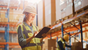 Woman in warehouse wearing hardhat looking at tablet.